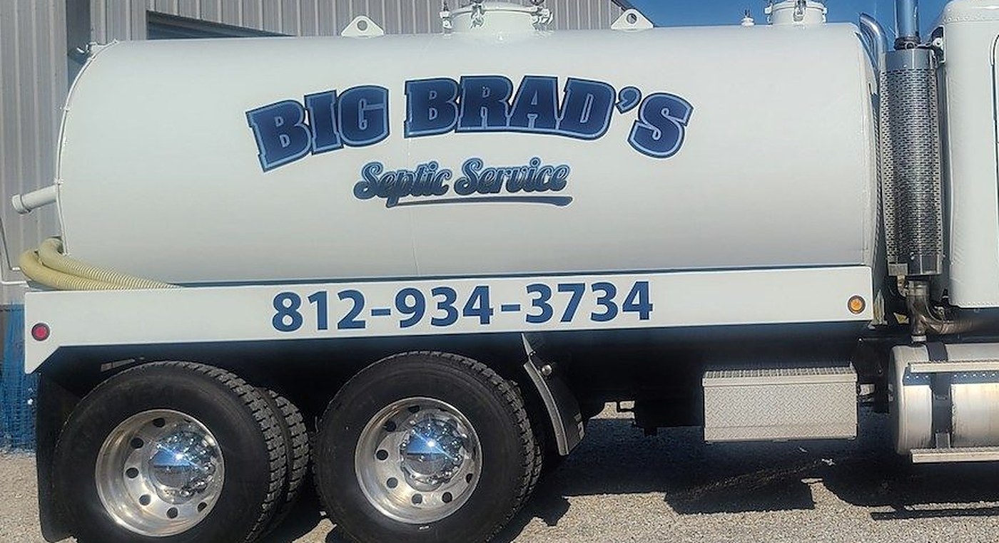 The white tanker truck parked on gravel beside a gray metal building, with Big Brad's Septic Service and the phone number 812-934-3734 lettered in blue along the side of the tank.